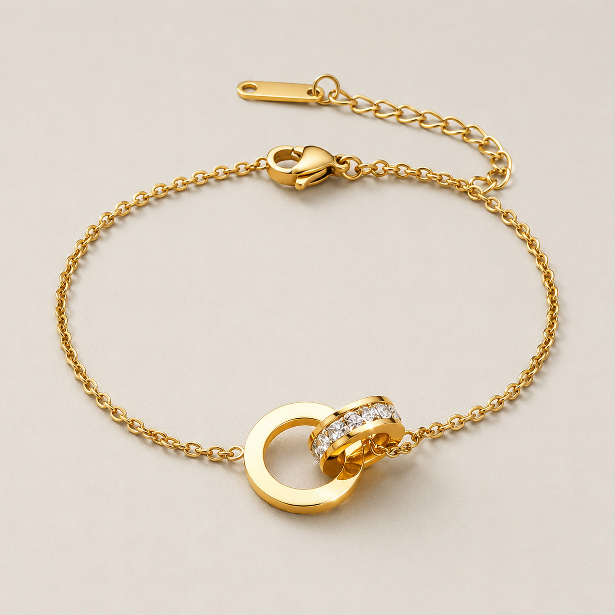 A gold chain bracelet with a double-ring pendant, one of which is adorned with small clear stones, displayed on a light beige background.