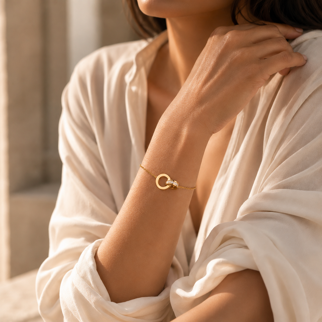 A close-up lifestyle shot of a woman wearing a delicate gold bracelet with a small interlocking rings design and a tiny clear crystal accent, styled with a flowing white blouse.