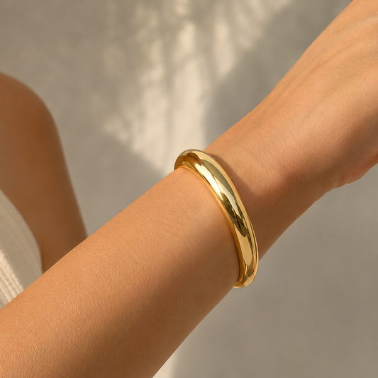 A polished gold bangle bracelet worn on a person's wrist, shown in a close-up shot against a soft, light gray background with subtle leaf shadows.